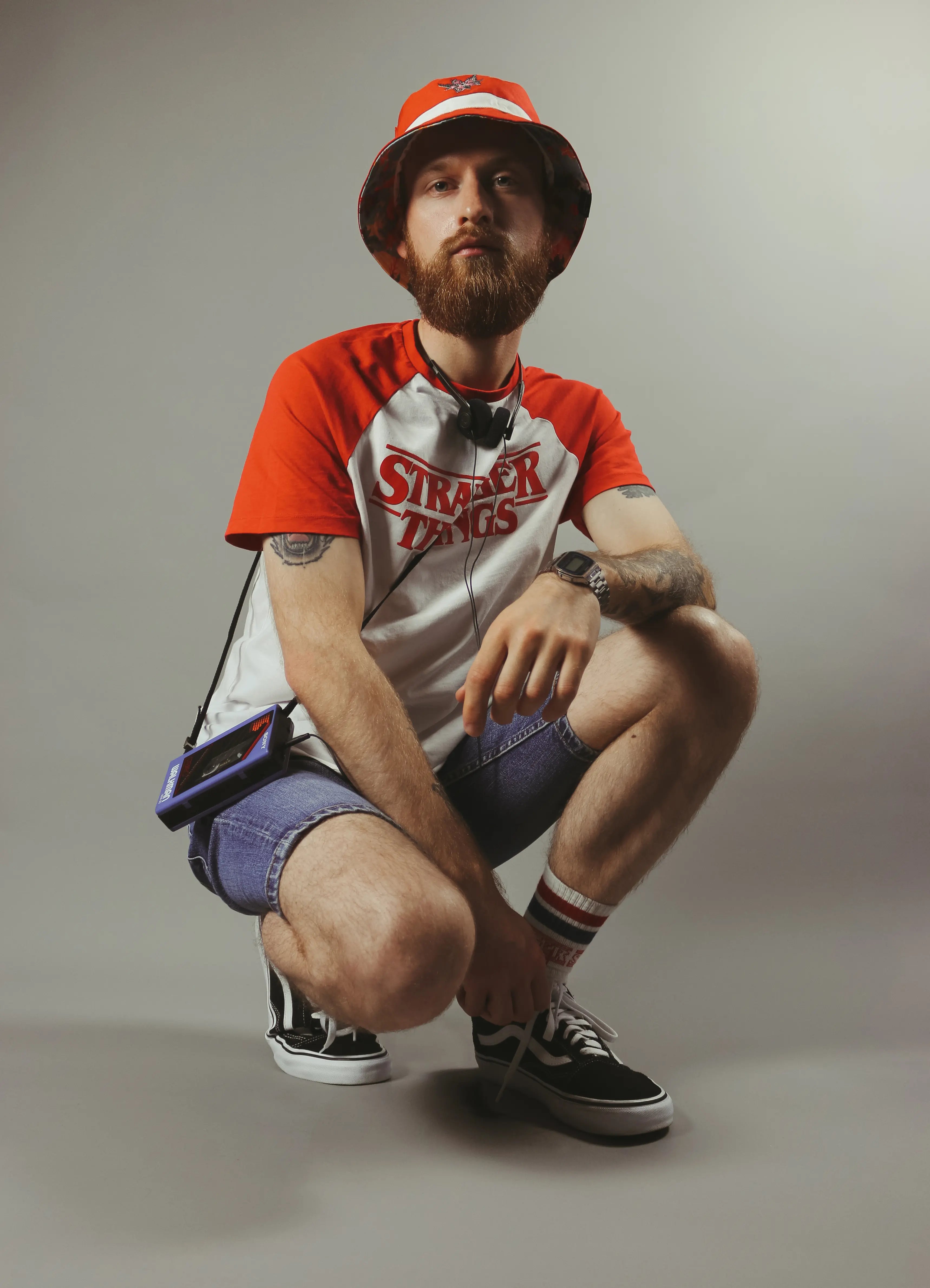 Mann in rotem Bucket-Hut, rotem T-Shirt mit weißem Einsatz und Aufdruck 'Stranger Things', Jeansshorts, Sneakers und Armbanduhr.