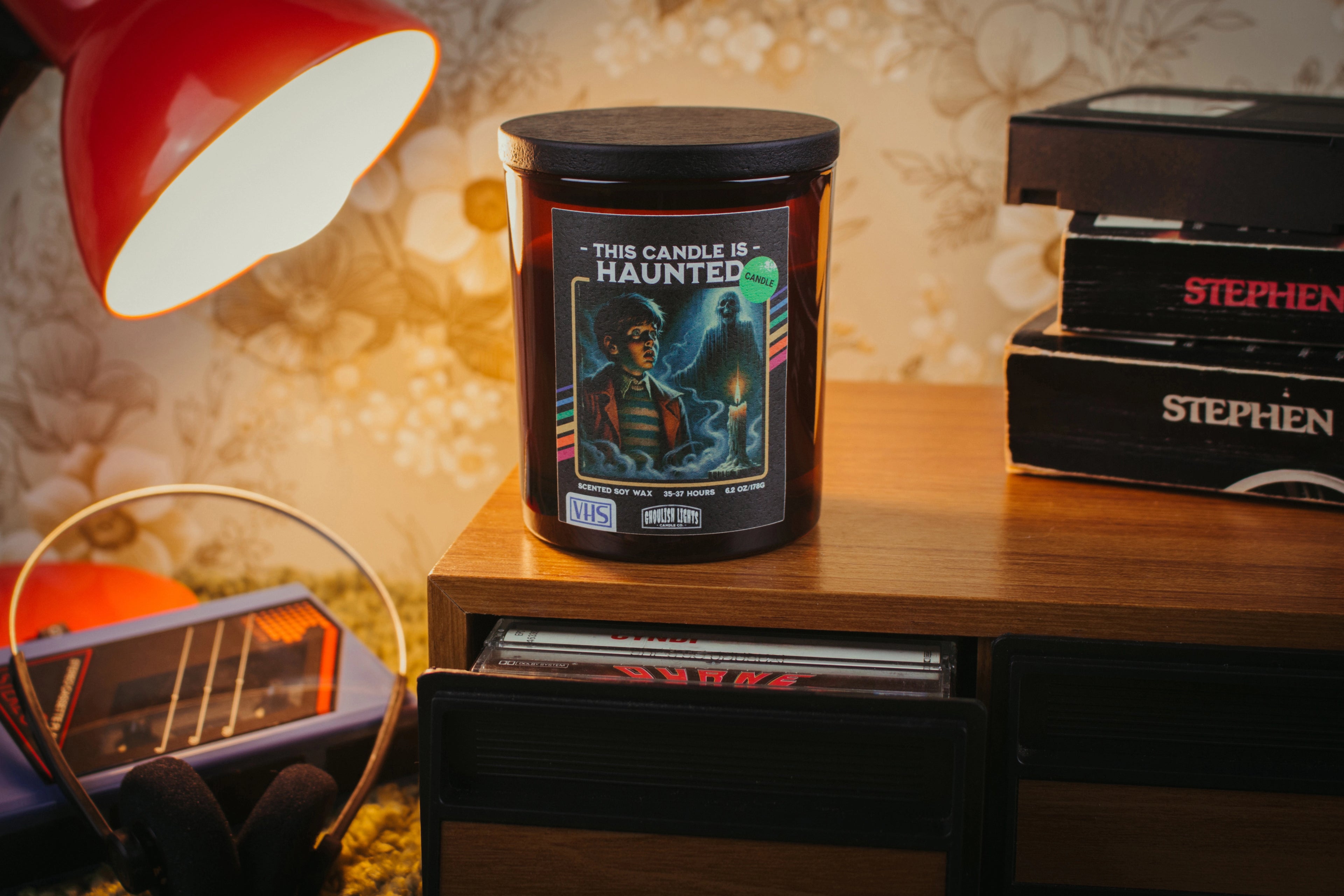 Handgemachte 80er-Horror-Kerze im VHS-Kassetten-Design auf Nussbaumholz-Deck vor vintage 80er-Jahre-Blumentapete