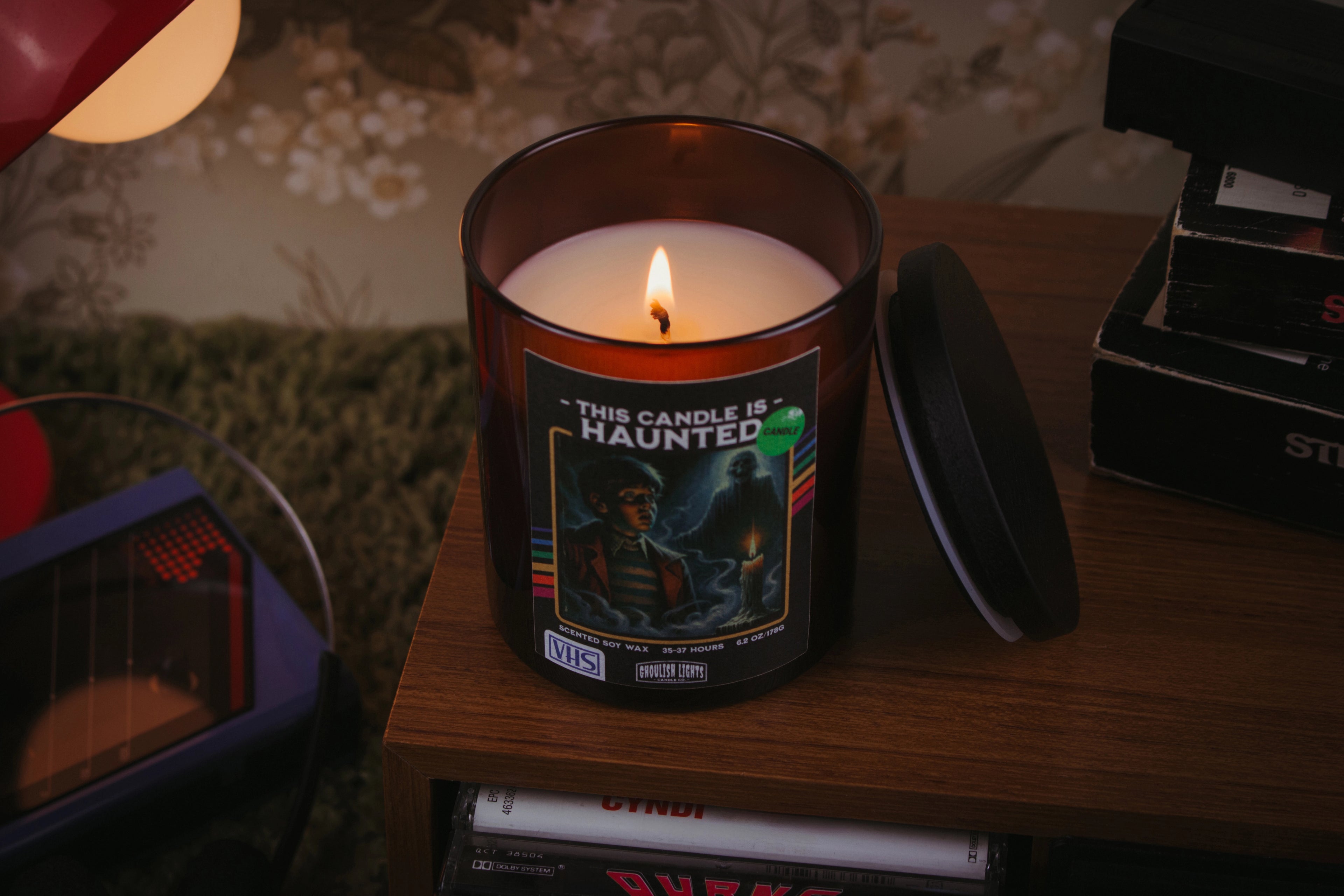 Handgemachte 80er-Horror-Kerze im VHS-Kassetten-Design auf Nussbaumholz-Deck vor vintage 80er-Jahre-Blumentapete