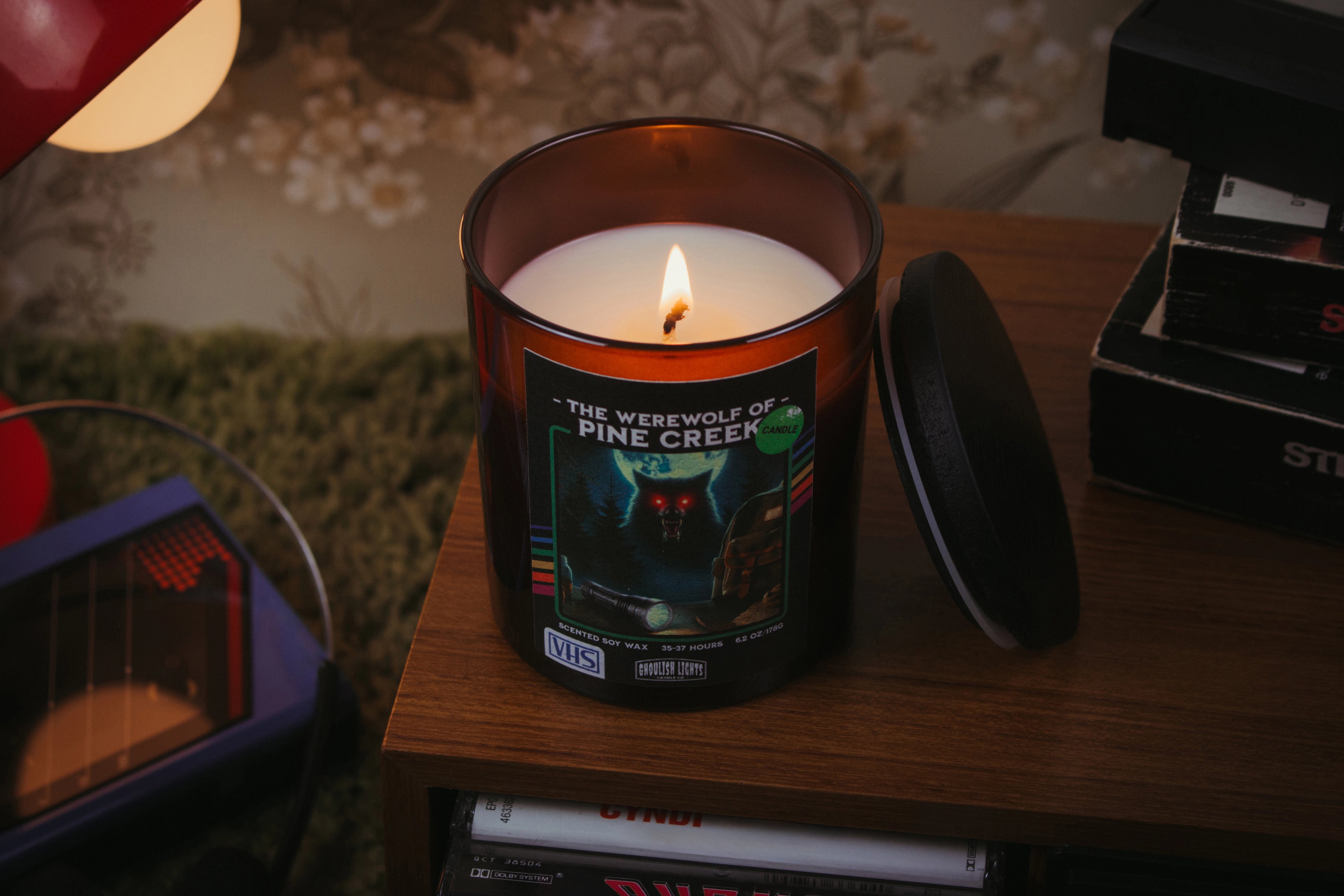 Handgemachte 80er-Horror-Kerze im VHS-Kassetten-Design auf Nussbaumholz-Deck vor vintage 80er-Jahre-Blumentapete