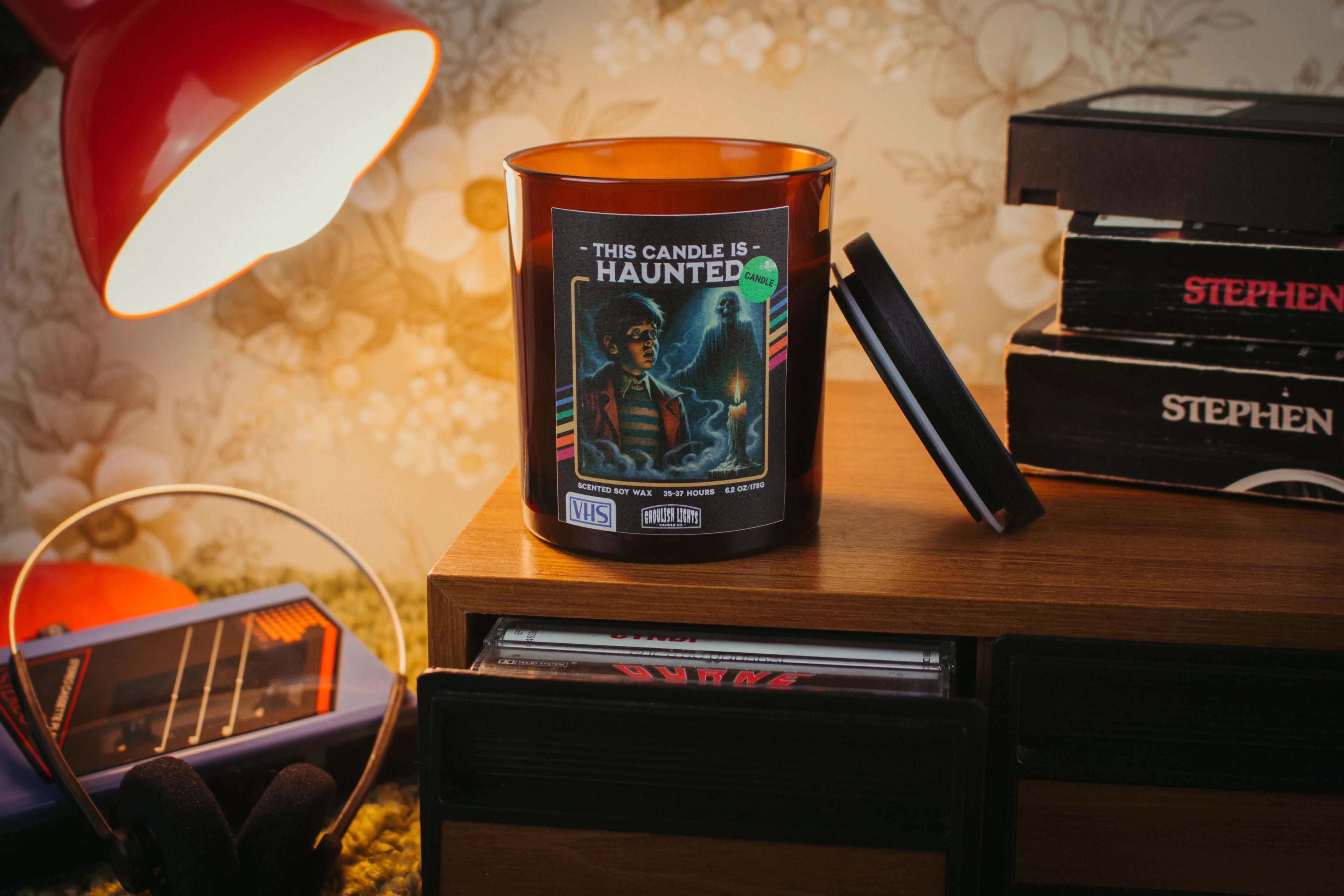 Handgemachte 80er-Horror-Kerze im VHS-Kassetten-Design auf Nussbaumholz-Deck vor vintage 80er-Jahre-Blumentapete
