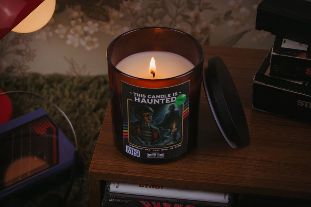 Handgemachte 80er-Horror-Kerze im VHS-Kassetten-Design auf Nussbaumholz-Deck vor vintage 80er-Jahre-Blumentapete