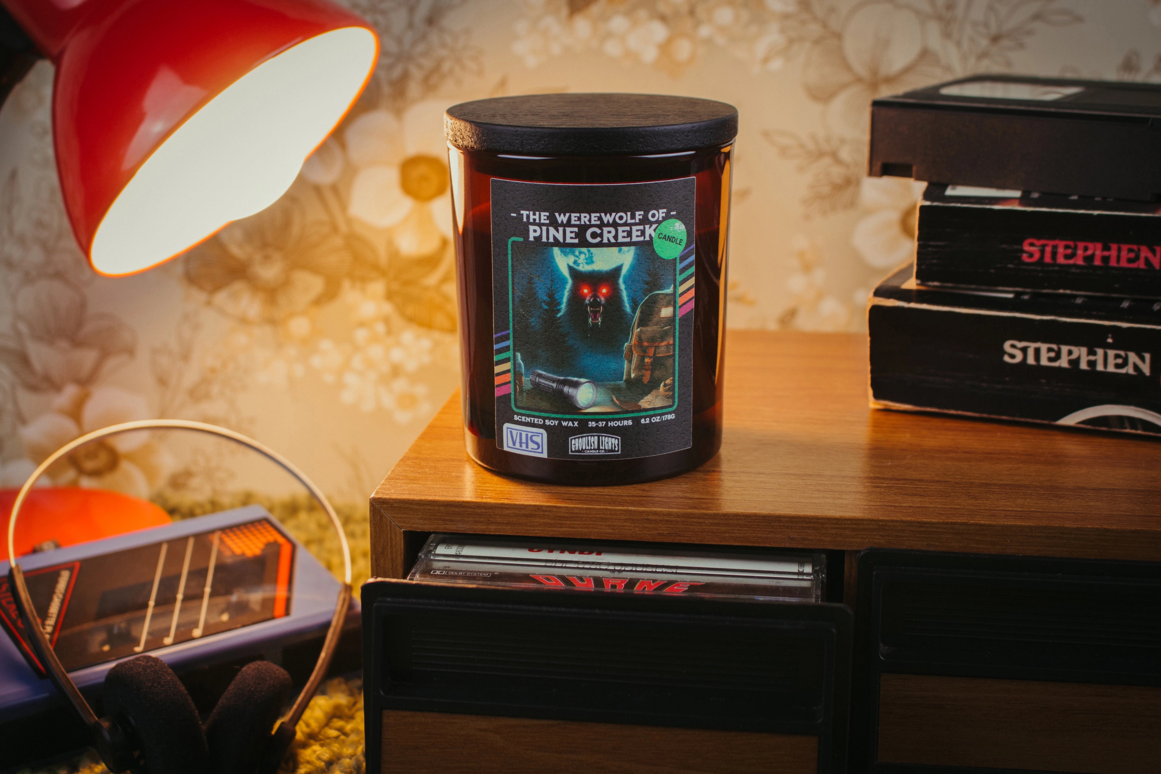 Handgemachte 80er-Horror-Kerze im VHS-Kassetten-Design auf Nussbaumholz-Deck vor vintage 80er-Jahre-Blumentapete
