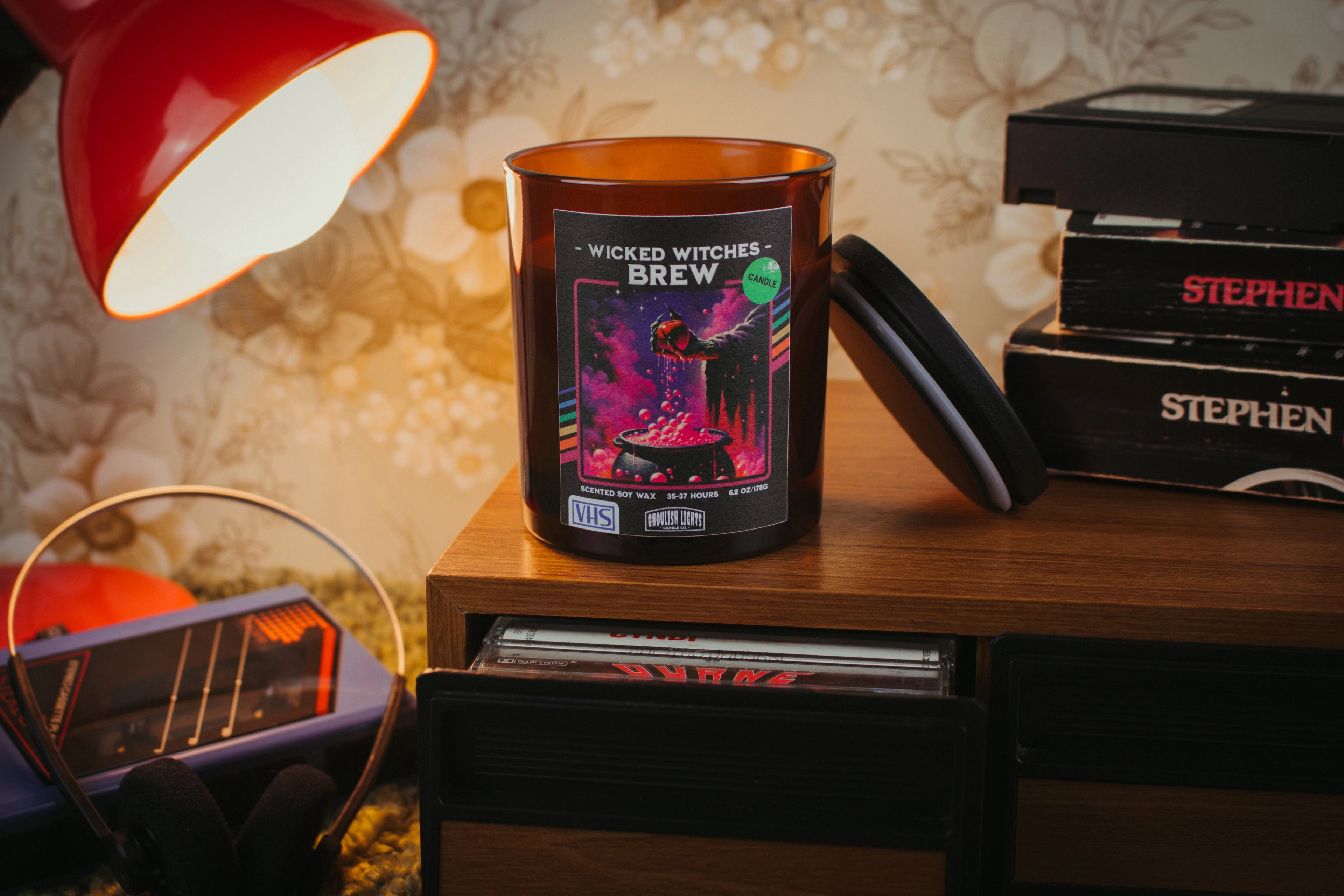 Handgemachte 80er-Horror-Kerze im VHS-Kassetten-Design auf Nussbaumholz-Deck vor vintage 80er-Jahre-Blumentapete