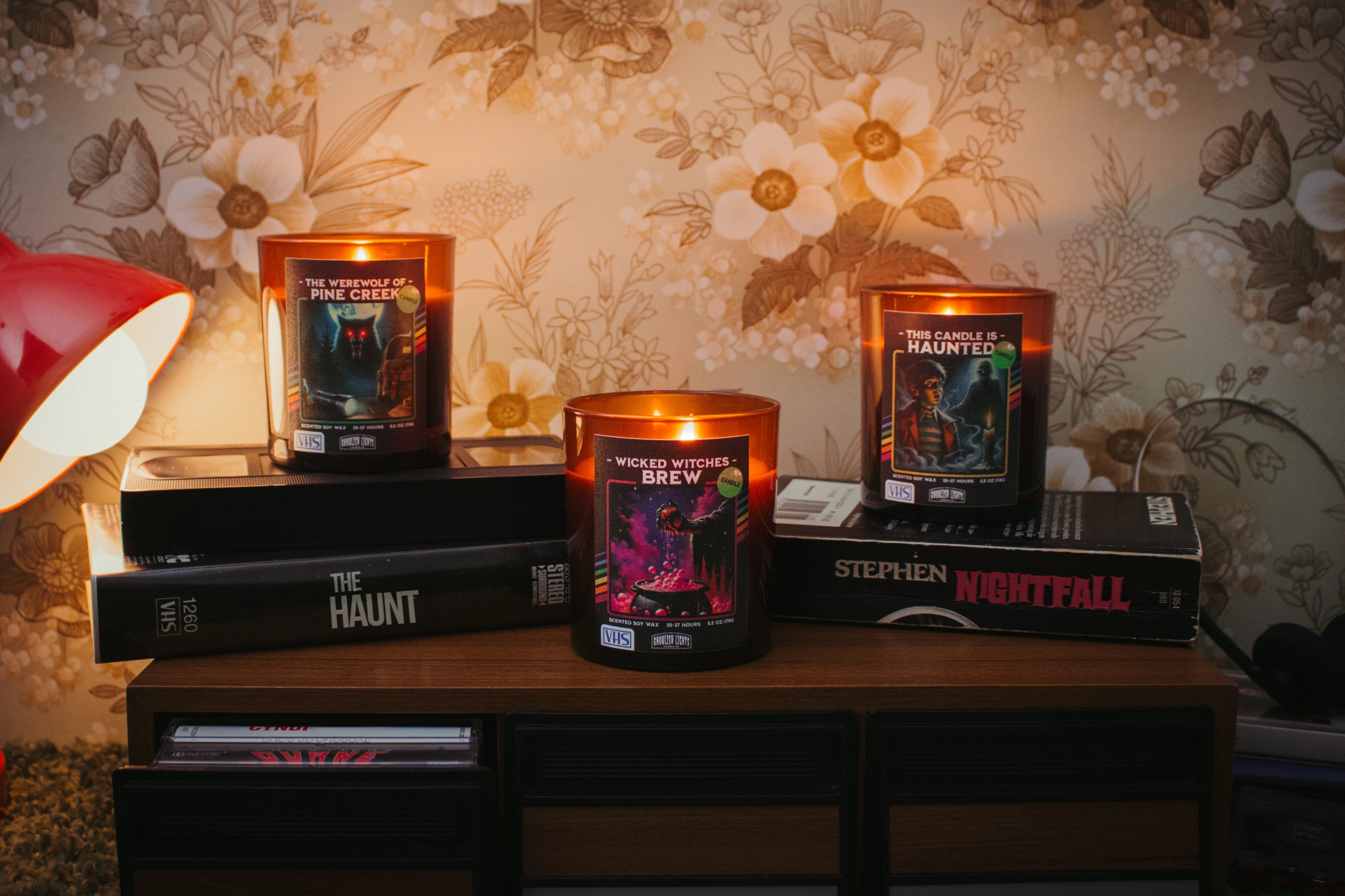 Drei handgemachte 80er-Horror-Kerzen im VHS-Kassetten-Design auf Nussbaumholz-Deck vor vintage 80er-Jahre-Blumentapete